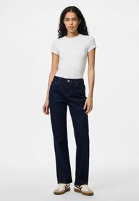 Balts īsām piedurknēm t-krekls apvienots ar tumši ziliem taisnās kājas džinsiem, ar parastām kabatām un pogu aizdari; valkāts ar bēšiem sporta apaviem.