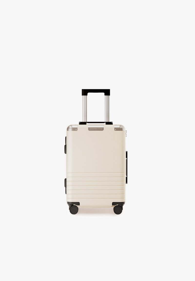 Hartschalenkoffer in Creme mit gerippter Textur. Verfügt über einen teleskopierbaren Griff, schwarze Akzente und vier schwarze Rollen für die Mobilität.