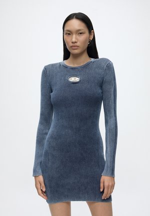 Jeune femme aux longs cheveux noirs portant une robe bleue côtelée ajustée à manches longues avec un détail métallique ovale sur la poitrine.