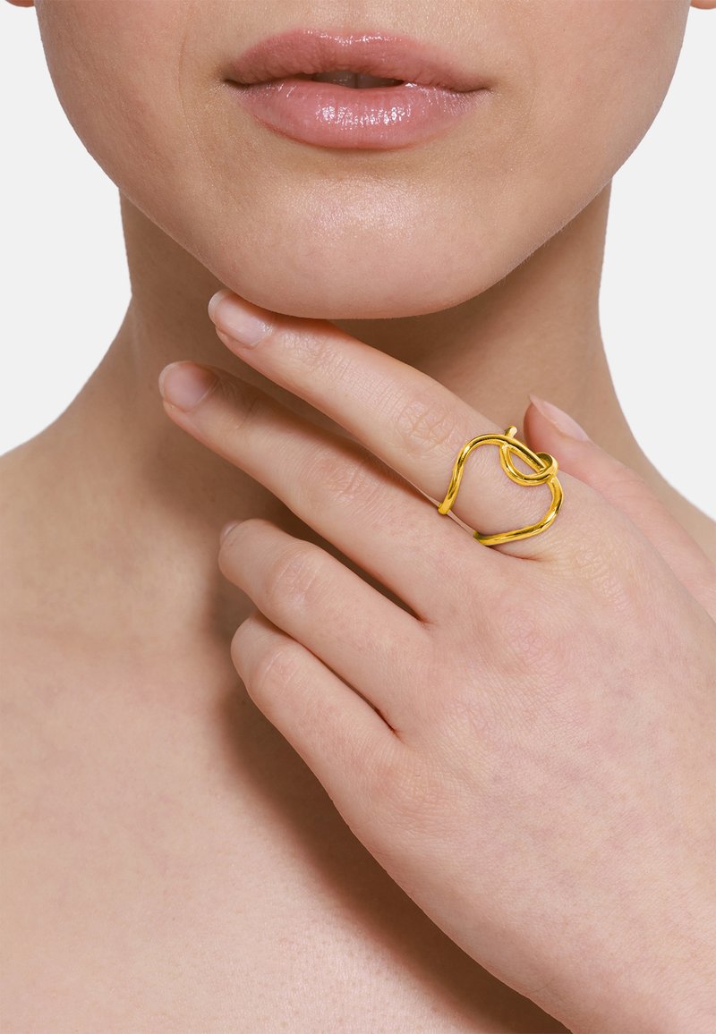 Bague dorée avec un design tordu, présentant des formes lisses et arrondies. Portée à l'index, mise en valeur sur un fond neutre.