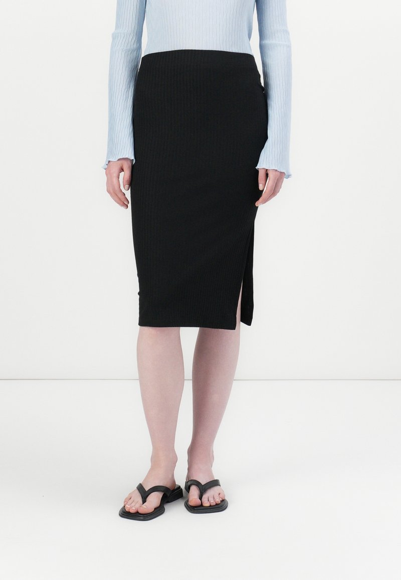 Femme portant une jupe midi noire côtelée avec une fente latérale, un haut à manches longues bleu clair et des sandales plates noires, debout sur un sol blanc.