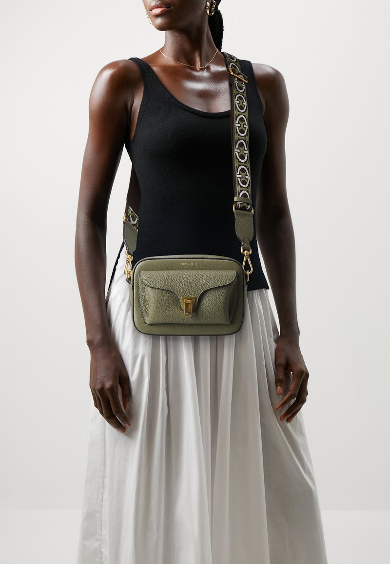 Borsa a tracolla rettangolare verde oliva in pelle texturizzata, con accessori dorati e tracolla regolabile a fantasia, indossata su una blusa nera e una gonna bianca.