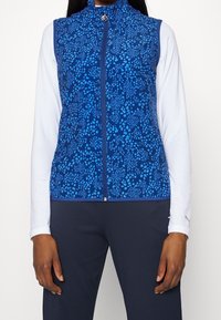 Blåmönstrad zip-up väst över vit långärmad skjorta, kombinerat med mörka rippade byxor, bärs av en person som står med armarna avslappnade.