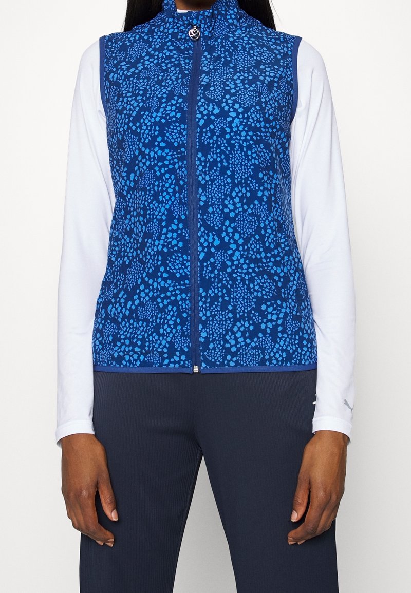 Blåmönstrad zip-up väst över vit långärmad skjorta, kombinerat med mörka rippade byxor, bärs av en person som står med armarna avslappnade.