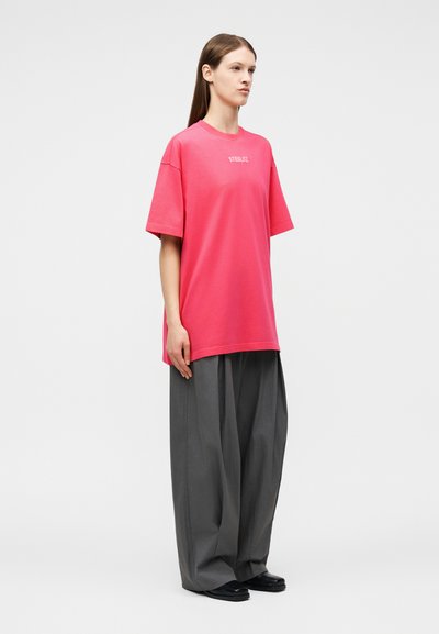 Νεαρή γυναίκα στέκεται πλευρικά φορώντας ένα μεγάλο φωτεινό ροζ μπλουζάκι με λευκό κείμενο και φαρδιά γκρι παντελόνια με μαύρα παπούτσια.