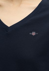 Gros plan d’un t-shirt bleu marine à col en V avec un petit logo GANT brodé représentant un blason avec des étoiles et des rayures sur la poitrine.