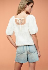Weiße, strukturierte Bluse mit gepufften Ärmeln und offenem Rücken, mit einer Schleife gebunden. Kombiniert mit ausgefransten Denim-Shorts. Der Hintergrund ist in einem sanften Orange.