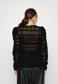 Femme aux cheveux foncés attachés en queue de cheval, portant un haut en dentelle noire transparente à manches bouffantes et un pantalon en cuir noir, vue de dos.