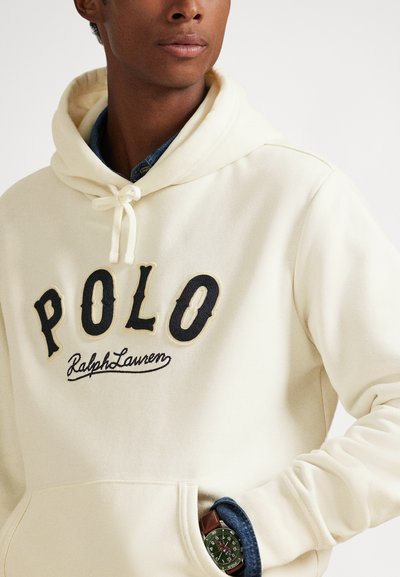 Asmuo, dėvintis kreminės spalvos gobtuvą su užrašu „POLO Ralph Lauren“ ir laikrodį su žaliu ciferblatu bei ruda odine apyranke kairėje riešo pusėje.