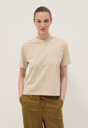 Vrouw met naar achteren gestreken haar, gekleed in een beige T-shirt met korte mouwen en olijfgroene broek met trekkoord, met handen in de zakken tegen een effen achtergrond.