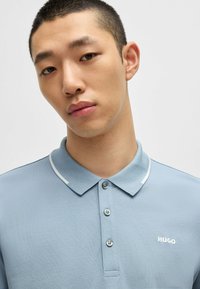 Lyseblå polo-shirt i et tekstureret stof, med tre knapper og et hvidt logo. Har en kontrasterende hvid kravekant.