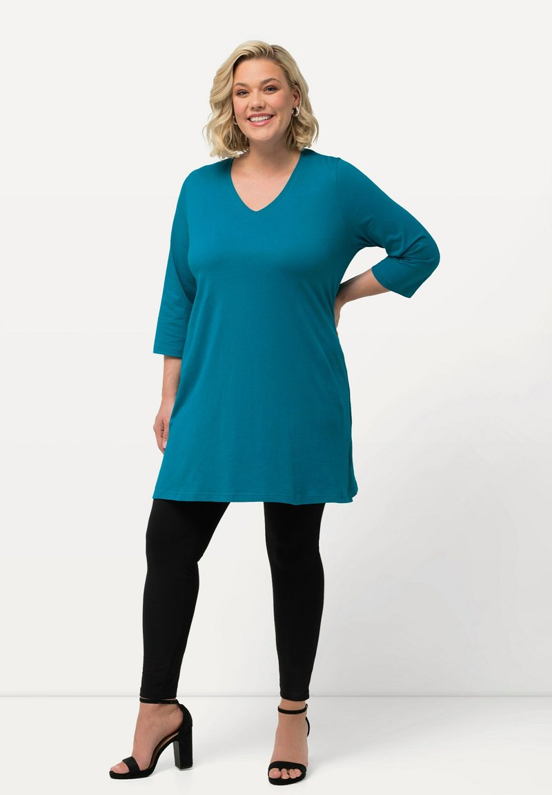 Tealfärgad tunika med V-ringning och trekvartsärmar, kombinerad med svarta leggings och högklackade sandaler. Slät tyg, avslappnad passform.