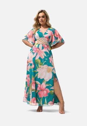 Kvinde iført teal blomsterprint slå-om-kjole med flæseærmer og sideslids, stående med hænderne på hoften og beige højhælede sandaler.