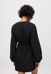 Čierne šaty s textúrovanou tkaninou, s užším pásom, dlhými puff rukávmi a okrúhlym výstrihom, strihané nad kolená. Predstavený pohľad zozadu.