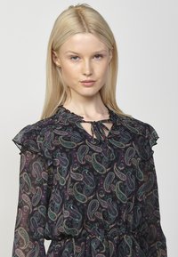 Tamsi gėlėta bluzonė su paisley raštu žalia, purpuru ir auksu. Sukurta su raukšlėmis pečių srityje ir kaklo rišimu. Lengvas, permatomas audinys.