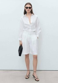 Weißes Leinenhemd mit tiefem V-Ausschnitt, plissierte weiße Shorts, schwarze geflochtene Clutch und schwarze Sandalen mit Riemchen und Absatz. Weiche Textur durchgehend.