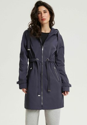 Kobieta z falującymi ciemnymi włosami, ubrana w granatowy płaszcz z kapturem na zamek z regulowanym paskiem w talii i szare spodnie, stojąca na jednolitym jasnym tle.