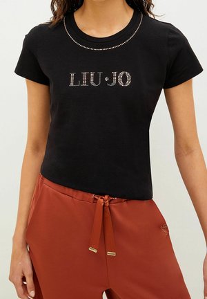 Kobieta ubrana w czarną koszulkę z krótkim rękawem z logo "LIU·JO" z cyrkoniami oraz rdzawo zabarwione spodnie z troczkami.