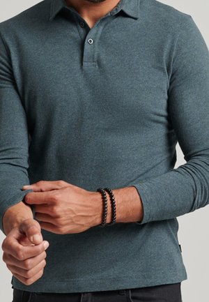 Long-sleeve polo shirt in dark teal, made of soft, textured fabric. Features a collar, two buttons, and visible cuffs. Black bead bracelets on wrist.
