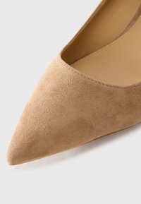 Close-up da ponta afunilada de um sapato de salto alto em camurça bege, com forro interior suave, sobre um fundo branco.