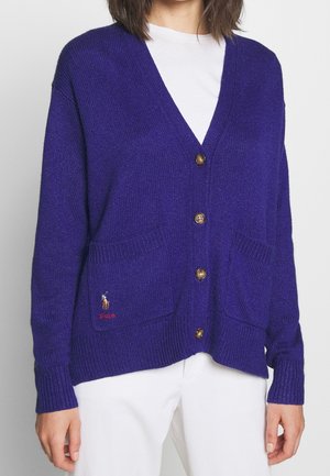 Frau trägt eine violette, geknöpfte Strickjacke mit vorderen Taschen und Polo-Logo auf der linken Tasche über einem weißen Hemd und weißen Hosen.