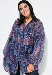 Mujer sonriente con cabello oscuro que lleva pendientes de aro y una blusa de botones transparente de patrones en azul, rojo y blanco con mangas amplias.