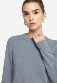 Maglione lavorato a maglia blu chiaro con scollatura tonda, polsini a coste e una texture spazzolata, indossato in modo casual. Design semplice con una finitura morbida.