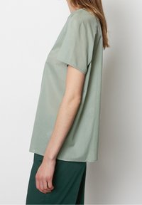 Persona che indossa una camicia a maniche corte verde chiaro e larga e pantaloni verde scuro, in piedi di lato su uno sfondo bianco semplice.