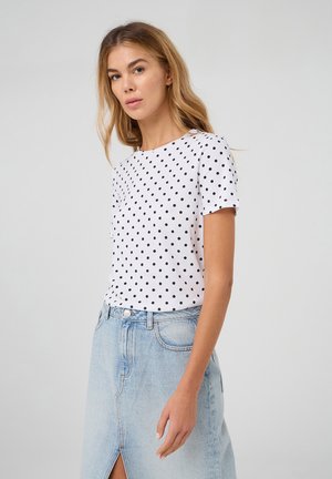 T-shirt bianco a maniche corte con pois neri; scollatura rotonda; abbinato a una gonna di jeans azzurri con uno spacco frontale.