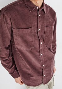 Camisa de pana en color malva con un cierre de botones en la parte delantera, dos bolsillos en el pecho y un cuello con solapas. Textura suave con acanalado vertical.