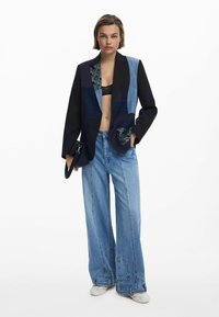 Patchwork blazer in dark blue, black, and denim with floral accents; paired with wide-leg blue jeans featuring detailed stitching.