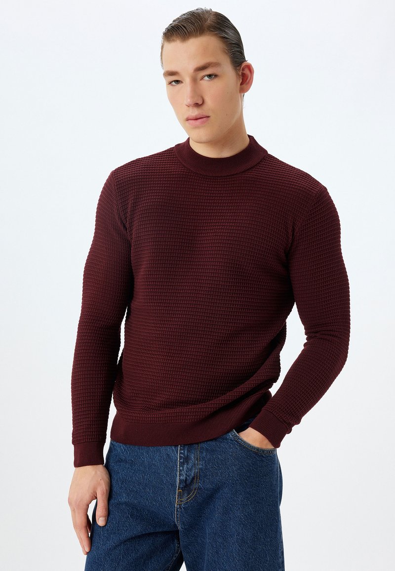 Maglione lavorato a maglia bordeaux con colletto alto, motivo a trama e polsini a coste. Abbinato a jeans in denim blu.
