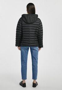 Czarna kurtka puchowa z poziomym pikowaniem, z kapturem, w połączeniu z niebieskimi dżinsami i czarnym obuwiem.