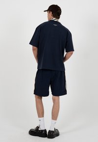 Homme portant un t-shirt bleu marine oversize et un short avec des bandes jaunes sur les côtés, des chaussettes blanches, des chaussures noires et une casquette marron foncé, debout, de dos.