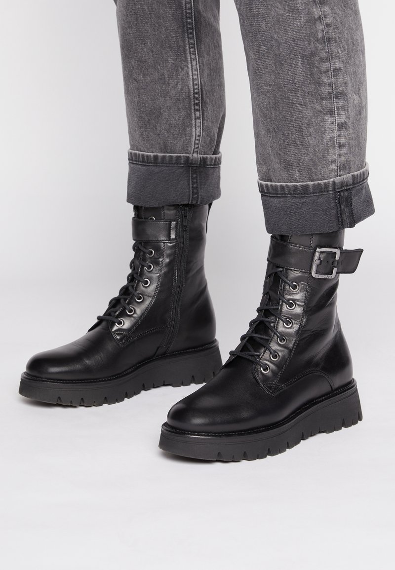 Bottines en cuir noir avec semelle épaisse, lacets à l'avant et fermeture éclair sur le côté. Présentent une sangle et une boucle au niveau de la cheville.