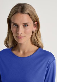 Blaues langärmeliges Oberteil mit Rundhalsausschnitt, weichem Material und lässiger Passform. Das Model trägt dezente Creolen und ein natürliches Make-up.