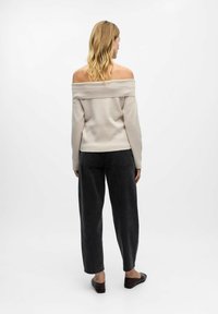 Bež pulover z nizkimi rameni, izdelan iz mehkega materiala, v kombinaciji s temnosivimi hlačami visokega pasu in ravnimi čevlji, prikazan zadaj.