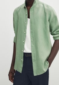 Ljusgrön linneskjorta med mandarin-krage, knäppning framtill och uppkavlade ärmar, bärs över en vit linne och mörk midjeresår.