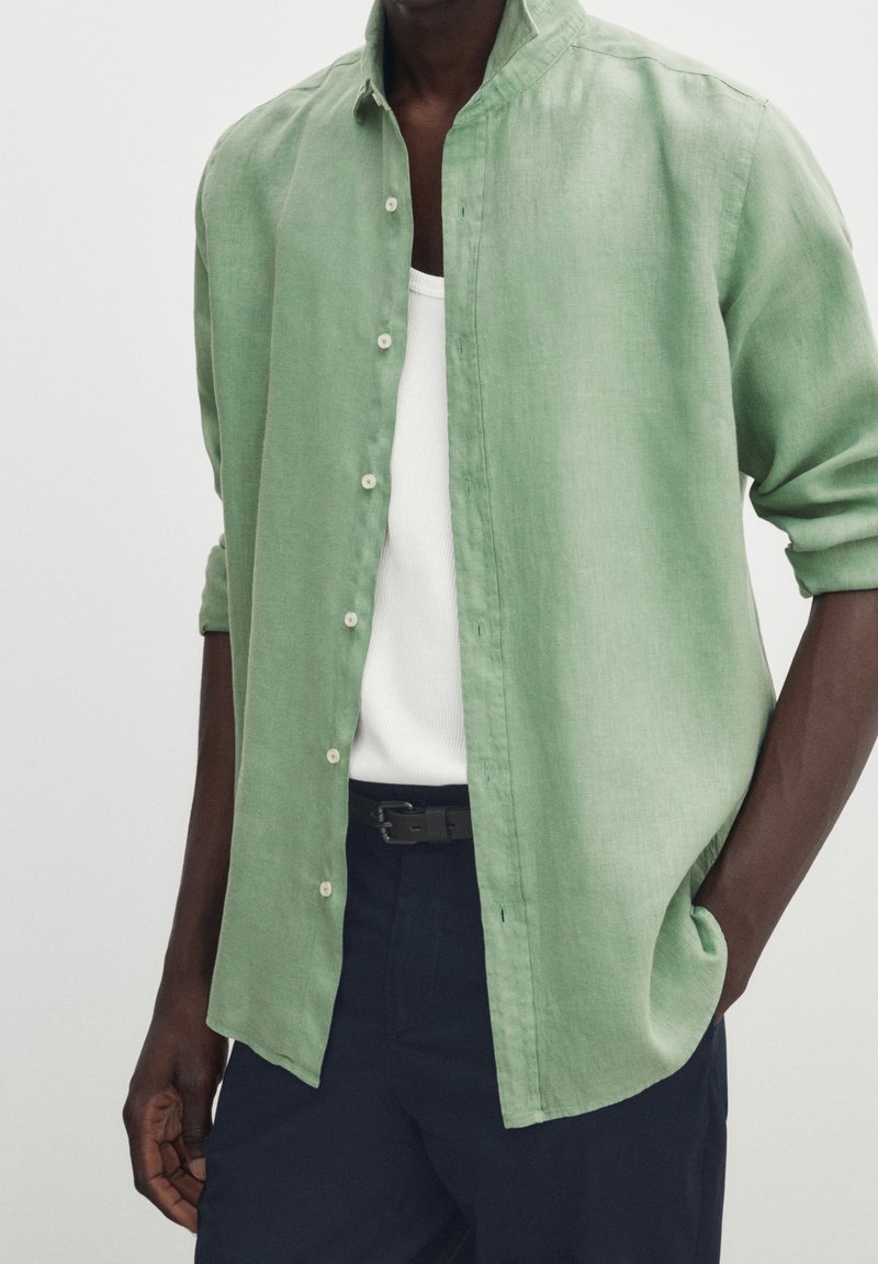 Ljusgrön linneskjorta med mandarin-krage, knäppning framtill och uppkavlade ärmar, bärs över en vit linne och mörk midjeresår.