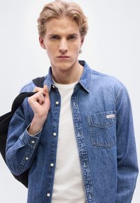 Πουκάμισο από τζιν σε ανοιχτό μπλε με κούμπωμα με κουμπιά, τσέπη στο στήθος και λογότυπο. Φορεμένο πάνω από ένα λευκό t-shirt, το μοντέλο κρατά μια μαύρη τσάντα.