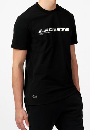 Mężczyzna ubrany w czarną koszulkę Lacoste z wyraźnym białym napisem logo na piersi i małym emblematem krokodyla przy krawędzi, stoi z jedną ręką w kieszeni.