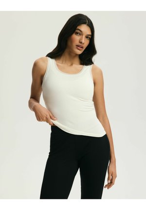 Kobieta o długich ciemnych włosach, ubrana w biały bezrękawnik i czarne spodnie, lekko podciągająca bluzkę z boku.