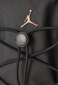 Juodas tekstūruotas paviršius su rožinės aukso Jumpman logotipu virš juodo apvalaus virvės užrakto, laikantį kryžmines juodas elastines virves.