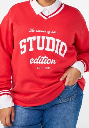 Personne portant un sweat-shirt rouge avec un texte blanc "The essence of now STUDIO edition EST. 1995" sur une chemise blanche à col et un jean bleu.