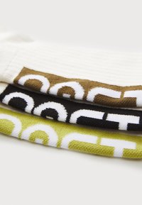 Trois paires de chaussettes en crème, vert olive et noir avec de grands motifs blancs. Chaque paire présente une texture côtelée et des designs graphiques distincts.