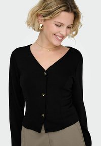 Ženska s kratkimi blond lasmi se smehlja in gleda navzdol, oblečena v črn kardigan z V-izrezom z gumbi v obliki srca in zlati obroči v ušesih.
