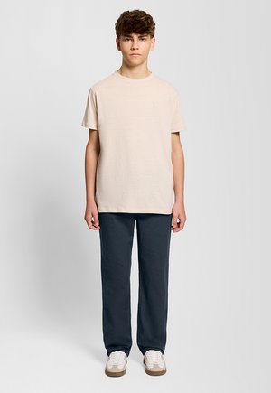 Ragazzo adolescente in piedi contro uno sfondo semplice, indossa una maglietta beige, pantaloni scuri e sneaker bianche, rivolto in avanti.