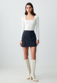 Top lungo a maniche ribbed bianco con scollatura quadrata, abbinato a una mini gonna a righe blu scuro e stivali bianchi fino al ginocchio.