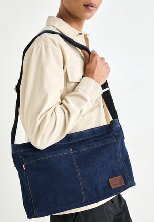 Personne portant une chemise beige tient un sac à bandoulière en jean bleu foncé avec un patch en cuir et une sangle réglable sur fond blanc.