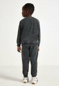 Tuta in maglia grigio scuro con scollatura rotonda a coste, polsini e vita elasticizzati; abbinato a sneakers bianche con accenti blu.